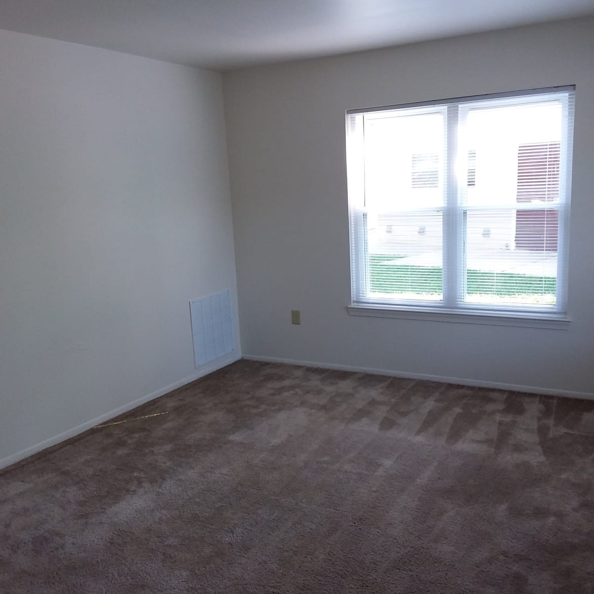 A carpeted bedroom at Indian Head Village with a wide double window, white blinds, and a return air grille in the wall