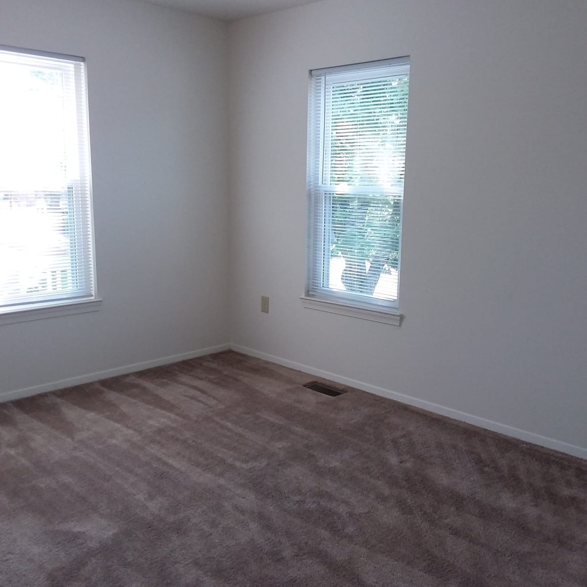 A corner bedroom at Indian Head Village with two windows on adjoining walls, white blinds, and wall-to-wall carpet