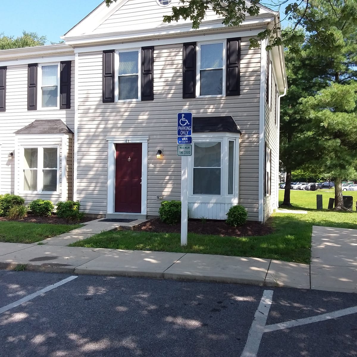 An end townhouse at Indian Head Village with light gray siding, black shutters, a dark red front door, and a bay window, with an accessible parking sign at the curb