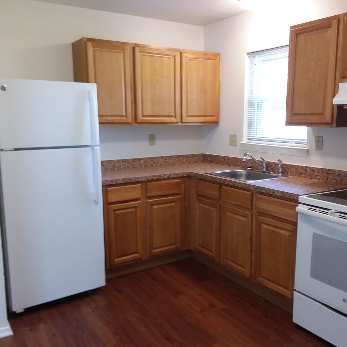 An L-shaped kitchen at Indian Head Village with oak cabinets, a white refrigerator and electric range, and a stainless sink under a window