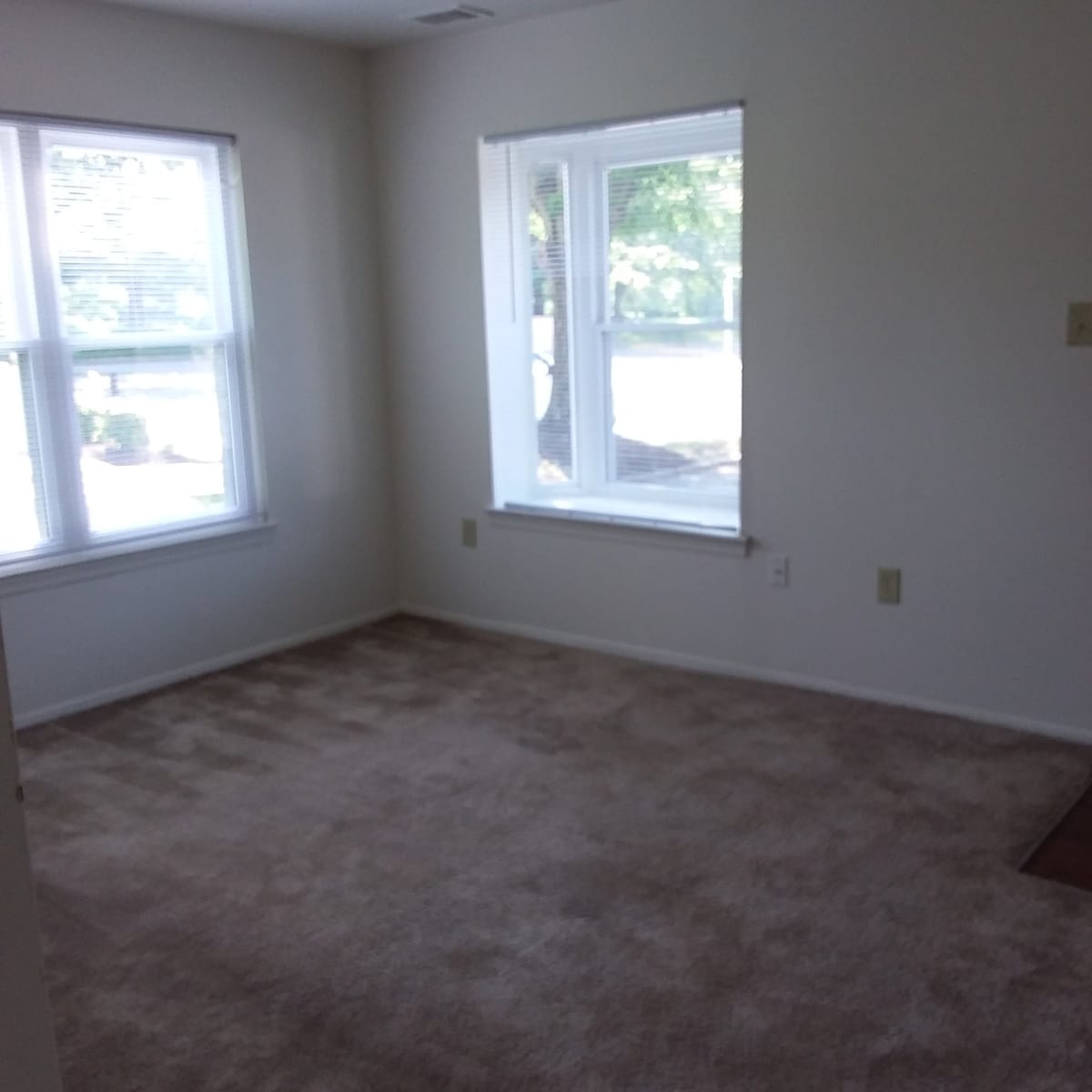 An empty carpeted living room at Indian Head Village with two windows and white blinds looking out to the front of the community