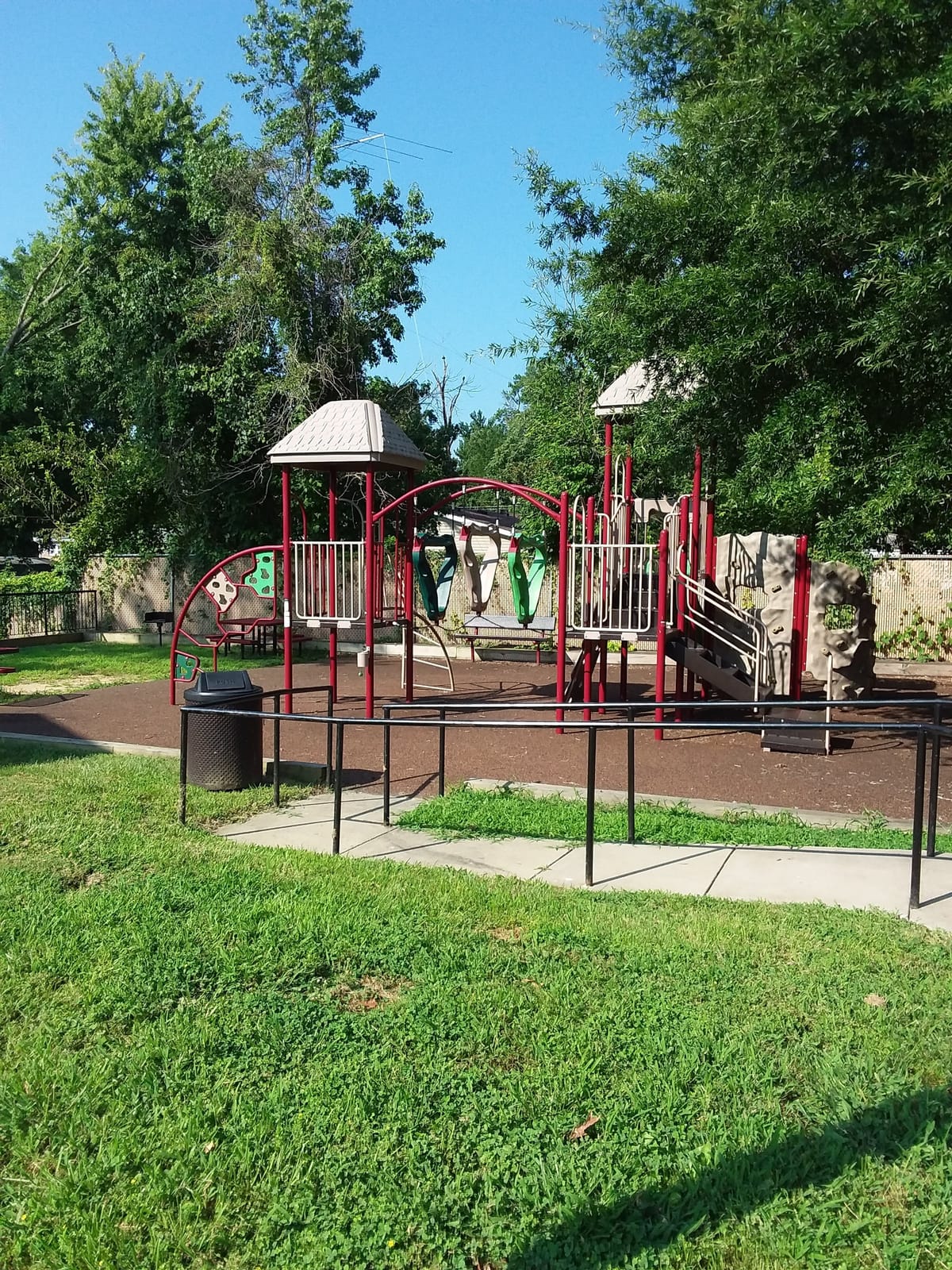 The playground at Indian Head Village, with a red and tan play structure, a climbing wall, and a picnic table on a rubber surface inside a railing