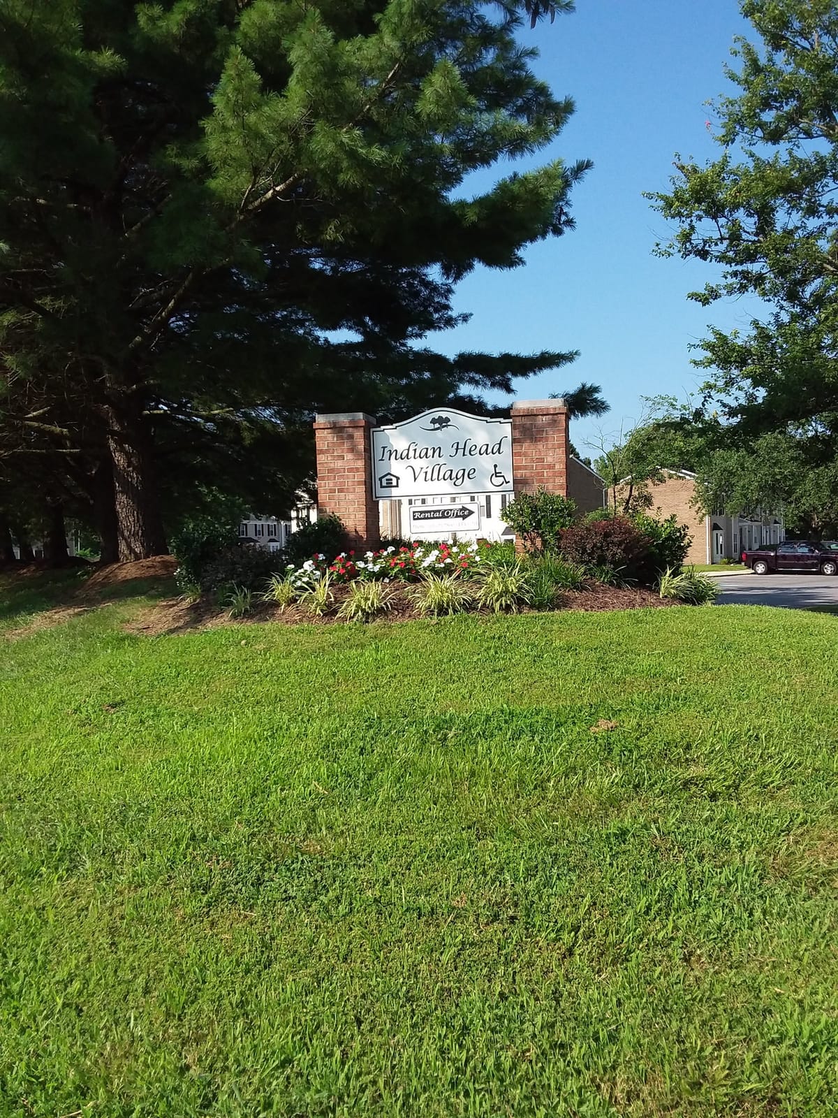 The Indian Head Village entrance sign on brick piers, set in a bed of red and white flowers beneath a tall pine